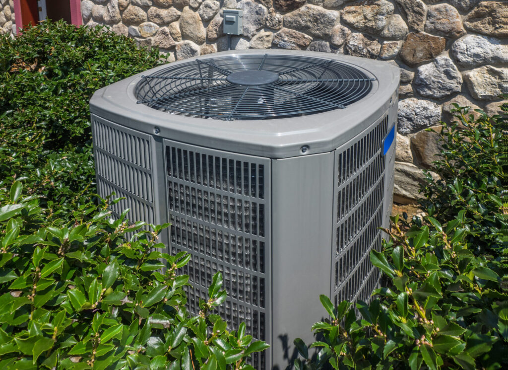 A photograph of a residential heat pump’s exterior unit. The unit is clean and well-maintained, tucked in behind some holly bushes.
